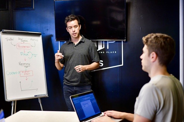 Ein Mann der neben einem Whiteboard steht und eine Präsentation hält. An einem Tisch vor ihm sitzt ein zweiter Mann mit einem Laptop, der zuhört.